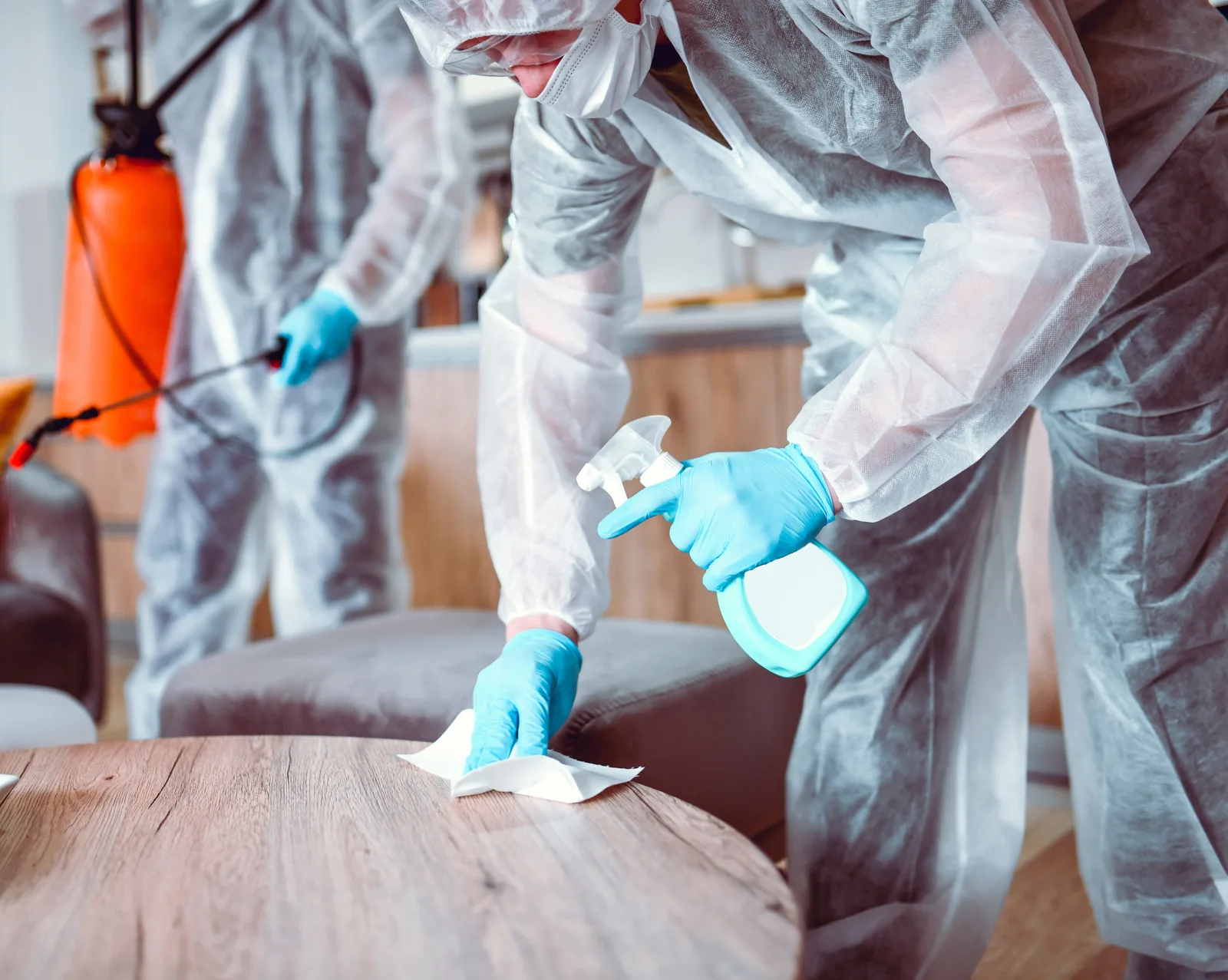 Two cleanup technicians in protective suits and gloves disinfecting a room